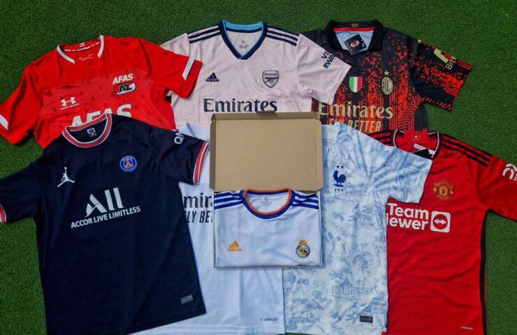 Collection de maillots de football soigneusement étalés sur un terrain artificiel vert, avec des maillots de clubs connus tels qu'Arsenal, Paris Saint-Germain, Real Madrid et Manchester United, chacun arborant ses sponsors tels qu'Emirates et TeamViewer, avec une boîte mystère ouverte au milieu.