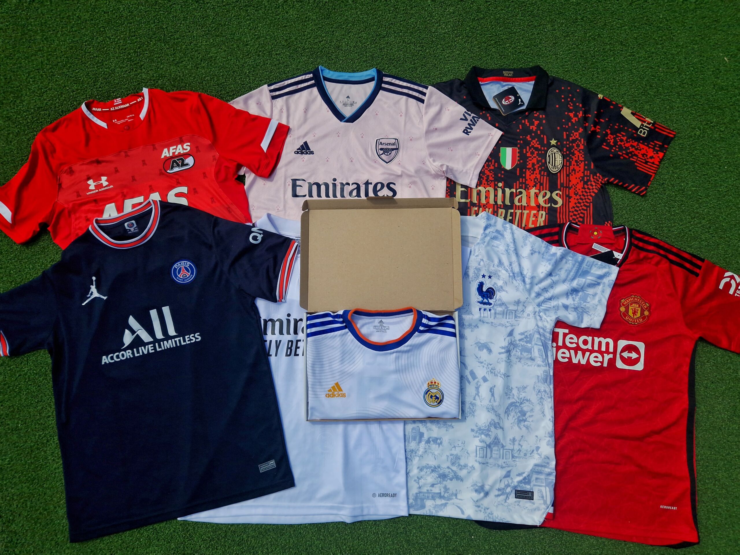 Collection of various football shirts carefully spread out on a green artificial pitch, with shirts from well-known clubs such as Arsenal, Paris Saint-Germain, Real Madrid, and Manchester United, each featuring their sponsors such as Emirates and TeamViewer, with an opened mystery box in the middle.