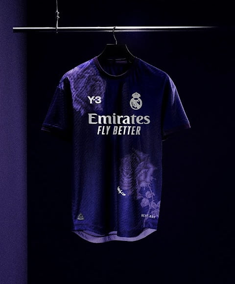 Maillot de football du Real Madrid suspendu en bleu marine avec le logo "Y-3" et le lettrage du sponsor "Emirates FLY BETTER", de subtils imprimés floraux et le logo du club, présentés sous un éclairage d'ambiance.